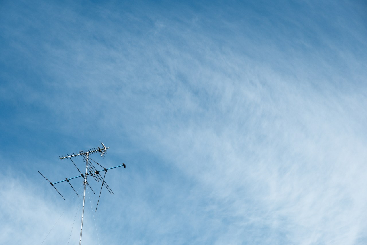 A metal television antenna is positioned in the lower-left corner of the frame against a vast, bright blue sky. Soft, wispy clouds stretch across the background, creating a sense of openness and clarity.